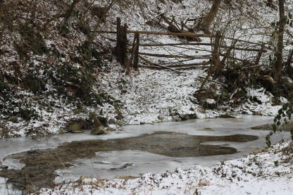 Zimsko je dan bez puno svjetla, tmurno vrijeme, potok je zaleđen, malo je snjiega. Uz potok trošne drvene ograde i šiblje.