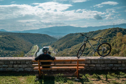 Biciklista sjedi i posmatra kanjon rijeke Une i grad Bihać sa starog grada Brekovice.