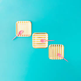 Tri papirna zlatno - bijela tanjura s tri ružičaste plastične kašike na plavoj pozadini.