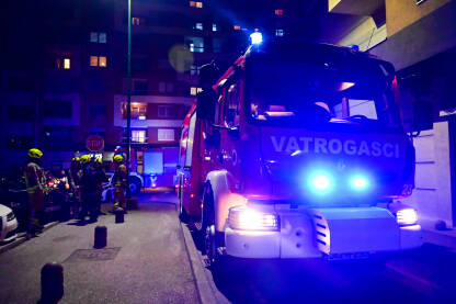 Vatrogasci uz vatrogasno vozilo sa upaljenim plavim svjetlima na intervenciji u gradu. Vatrogasna jedinica gasi požar u zgradi.
