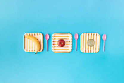 Piknik scena s tri papirna tanjura i tri plastične kašike, bananom, jabukom i konzervom na plavoj pozadini.