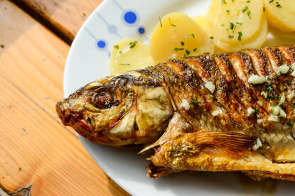 Riba grilovana na žaru u restoranu. Ukusna i slasna riba s krumpirom, limunom i češnjakom na tanjiru na drvenom stolu. Hrana.