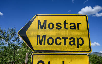 Žuti saobraćajni znak koji pokazuje lijevo s nazivom grada "Mostar" ispisanim i latinicom i ćirilicom. Mostar je grad u Bosni i Hercegovini.