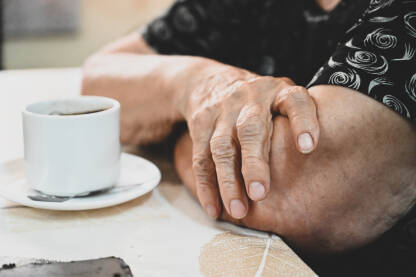 Ruke starije žene s bijelom šalicom kafe na stolu. Naborane ruke starice.