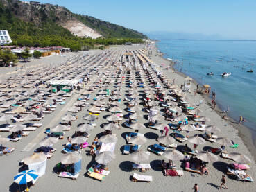 Mnogo ljudi na pješčanoj plaži i u moru. Suncobrani i ležaljke Mnogo ljudi na pješčanoj plaži i u moru. Suncobrani i ležaljke na obali uz more, snimak dronom. Jadransko more, Shengjin, Albanija. Turisti na plaži.