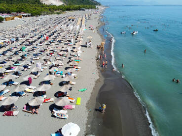 Mnogo ljudi na pješčanoj plaži i u moru. Suncobrani i ležaljke Mnogo ljudi na pješčanoj plaži i u moru. Suncobrani i ležaljke na obali uz more, snimak dronom. Jadransko more, Shengjin, Albanija. Turisti na plaži.