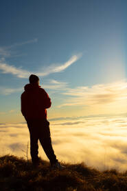 Planinar stoji na vrhu brda i posmatra sunce iznad kotline ispunjene maglom.