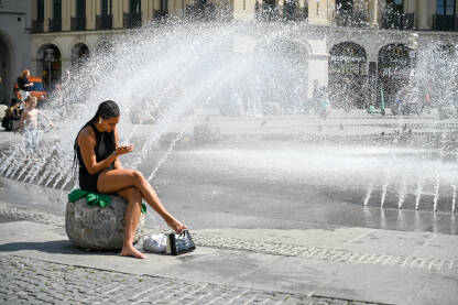 Mlada žena se osvježava kraj fontane. Djevojka se osvježava kraj vode u vrućem ljetnom danu. Toplotni talas u gradu.
