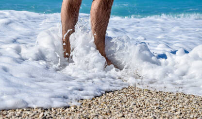Muškarac stoji u moru. Noge muškarca zapljuskuju morski talasi i pjena.