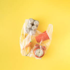 Plastična providna vrećica za kupovinu s katronskom ambalažom za jaja, mrkvom, bananom, satom i plastičnom čašom na žutoj pozadini.