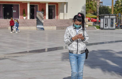 Djevojka stoji na ulici sa maskom na licu i drži telefon.