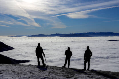 Tri planinara, siluete, stoje na ivici posmatrajući dolinu u magli