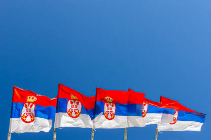 Zastave republike Srbije fotografisane nasuprot plavog neba, dosta praznog prostora.