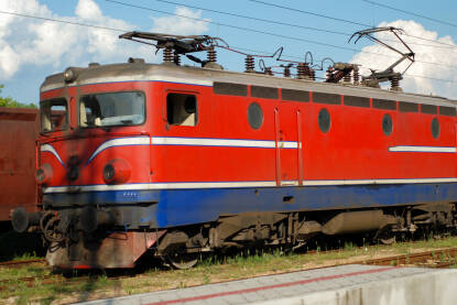 Crvena lokomotiva parkirana na peronu željezničke stanice, spremna za prevoz putnika. Krupni kadar jarkocrvene lokomotive na šinama na željezničkoj stanici. Putovanje vozom.