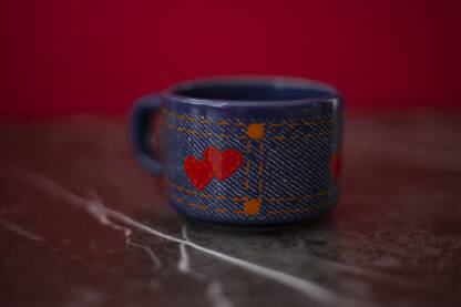 Plava solja za kafu sa detaljima u obliku rvenih srca i narandzastih linija, crvena pozadina i siva ploca na kojoj je solja