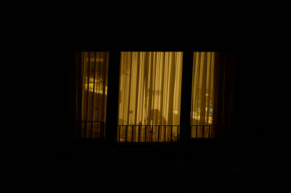 Osvjetljeni prozor tokom noći i žena koji sjedi za stolom. Silueta žene koja sjedi iza zavjese.