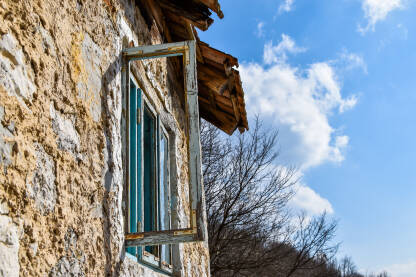 Stara, ruševna kuća od kamena, polusrušeni krov i drveni prozor bez stakla. Stara kuća u prirodi, tokom vedrog, proljetnog dana.