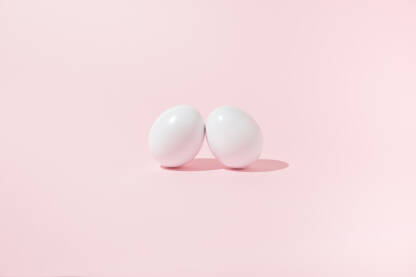 Dva bijela jaja naslonjena jedno na drugo na ružičastoj pozadini.