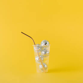 Plastična čaša za piće napunjena sjajnim disko kuglama i zlatnom slamkom na žutoj pozadini.