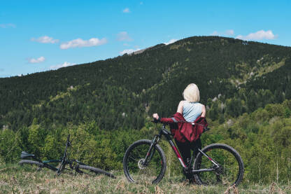 Biciklizam u prirodi. Djevojka naslonjena na bicikl sa pogledom na vrhove planina.