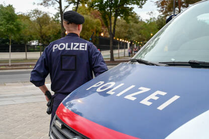 Policajac stoji uz policijski auto u Beču, Austrija. Policajac u uniformi patrolira ulicom. Simbol policije na vozilu i uniformi.