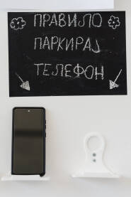 Ćirilični natpis "Pravilo parkiraj telefon" napisan kredom na crnom mat papiru, ispod natpisa na stalku ostavljen mobilni telefon.