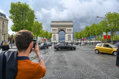 Pariz, Francuska: muškarac mobilnim telefonom fotografiše Arc de Triomphe u Parizu. Čuveni spomenik se nalazi na zapadnom kraju Champs-Élysées. Turisti istražuju Pariz.