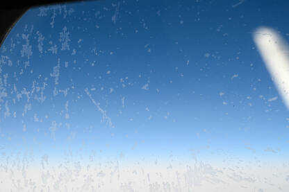 Smrznute kapljice kiše na prozoru aviona sa plavim nebom u pozadini. Pogled kroz avionski prozor tokom leta.  Ledene kapljice vode na prozoru aviona.