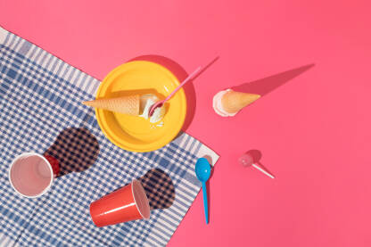 Piknik scena sa žutim plastičnim tanjurom, sladoledima, crvenim čašama i lizalicom na ružičastoj pozadini.