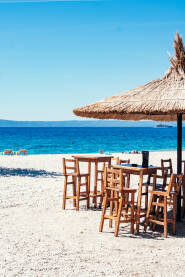 Šljunčana plaža sa suncobranima od trske, ležaljkama, drvenim stolovima i stolicama i pogledom na ostrva Brač i Hvar. Makarska.