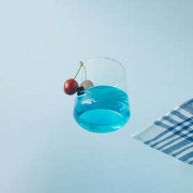 Čaša za piće s plavim napitkom i dvije svježe trešnje te stolnjakom sa strane na staklenoj površini.