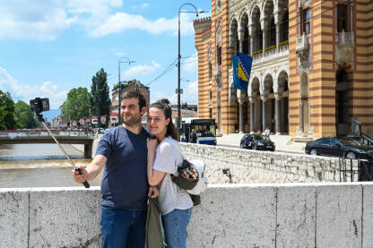 Turisti prave selfie ispred zgrade gradske Vijećnice u Sarajevu. Ljudi fotografišu mobilnim telefonima.