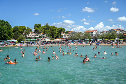 Grupa ljudi se sunča na plaži i kupa u Jadranskom moru. Turisti uživaju i zabavljaju se na plaži. Prepuna plaža. Turizam.