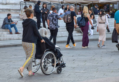 Žena gura invalidska kolica u kojima sjedi druga, starija žena. Osobe sa invaliditetom u javnom prostoru.