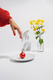 Ženska ruka koja poseže za crvenom ukrasnom kuglicom na bijelom tanjuru i buket žutog cvijeća.