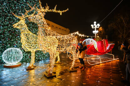 Ljudi prave fotografije u blizini okićenog drveta i osvijetljenih saonica Djeda Mraza. Okićeno drvce u centru grada noću. Šarene dekoracije.