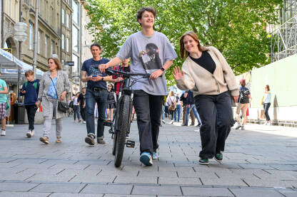 Minhen, Njemačka: Ljudi šetaju ulicom. Mlada djevojka maše prema kameri i mladić s biciklom. Minhen je popularna turistička destinacija u Bavarskoj.