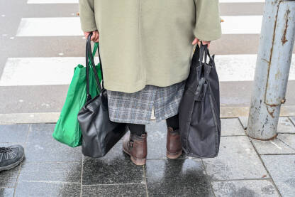 Starija žena sa torbama u ruci. Starica na trotoaru. Penzioneri idu u kupovinu.