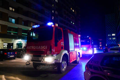 Vatrogasci uz vatrogasno vozilo sa upaljenim plavim svjetlima na intervenciji u gradu. Vatrogasna jedinica gasi požar u zgradi.