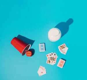 Igra pokera s kartama za igranje, crvenom plastičnom čašom, kockom i bijelom bocom za piće na plavoj pozadini.