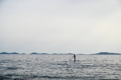 Silueta čovjeka koji surfa na dasci za veslanje. Veslanje po tihom moru tokom oblačnog dana.
