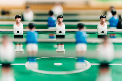 Fotografija prikazuje stolni fudbalski sto sa igračima u uniformama. Igrači nose plave i bele uniforme. Sto je zelene boje sa belim linijama koje označavaju igralište.