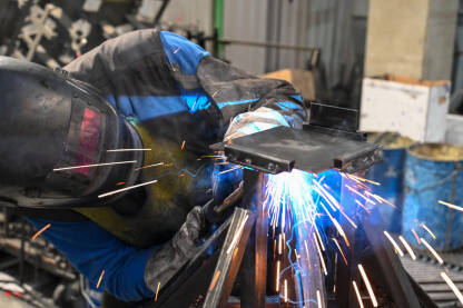 Industrijski radnik izvodi zavarivanje na metalnoj konstrukciji. Zavarivač radi u tvornici dok oko njega lete užarene iskre.