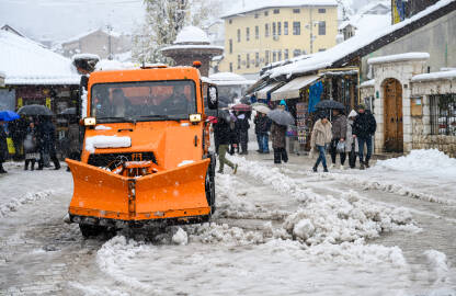 Narandžasta ralica čisti ulice Baščaršije tokom obilnih snježnih padavina, dok prolaznici prolaze sa kišobranima.