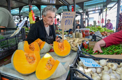 Starija žena prodaje voće i povrće na pijaci. Svježe organsko povrće na pijaci. Bundeve ili tikve na štandu.