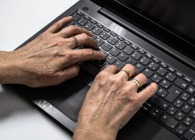 Starija ženska osoba kuca na tastaturi crnog lap-topa koji se nalazi na beloj pozadini. Stariji ljudi sve više uče da koriste računare.