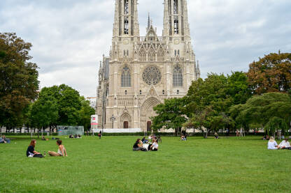 Votivkirche, neogotička crkva u Beču, Austrija. Mladi se druže na travi u parku. Ljudi sjede na zelenoj travi u parku.