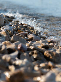 Mali morski talas zapljuskuje mokro kamenje na plaži tokom sunčanog ljetnog dana. Krupni plan mokrog namenja i morske vode.