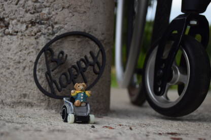 Igračka u invalidskim kolicima. Medo bez noge u kolicima, pored znaka "Budi srećan". U pozadini se vide točkići kolica za osobe sa invaliditetom.