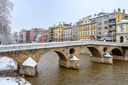 Zimski pogled na Latinsku ćupriju u Sarajevu, historični kameni most koji premošćuje rijeku Miljacku. Snijeg na lukovima mosta i okolnim zgradama stvara prepoznatljivu zimsku atmosferu Starog Grada.